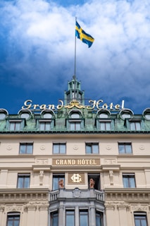 An elegant building with an ornate facade featuring the name 'Grand Hôtel.' The structure has multiple arched windows on the upper levels and a green-colored roof topped with a tall flagpole displaying a flag. The architectural design includes decorative elements and statues.