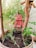 A small clay robot with a simple face and a heart symbol on its chest is placed amidst potted plants. The robot is crafted with a rectangular head and body, positioned between two wooden sticks. It is surrounded by green foliage and sits in dark, moist soil inside a round planter.