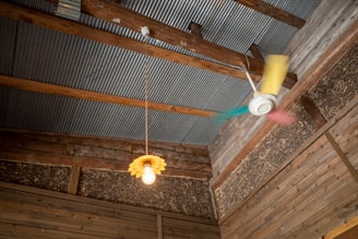 The interior of a rustic room with a corrugated metal ceiling and wooden beams. A vintage-style yellow pendant light with an exposed bulb hangs from the ceiling next to a colorful ceiling fan with blurred blades in motion. The walls are made of wood with visible straw insulation.