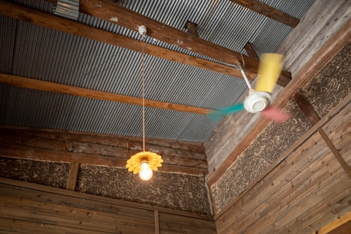 The interior of a rustic room with a corrugated metal ceiling and wooden beams. A vintage-style yellow pendant light with an exposed bulb hangs from the ceiling next to a colorful ceiling fan with blurred blades in motion. The walls are made of wood with visible straw insulation.