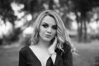 A woman with long curly hair gazes confidently at the camera, with striking, digitally enhanced green eyes that stand out against the black-and-white background. She wears a dark, textured top and a bracelet on her wrist. The blurred background consists of trees and foliage.
