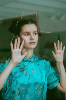 A person is standing with their hands slightly raised, wearing a teal-colored traditional Chinese dress with intricate patterns. Their hair is adorned with sticks styled in an artistic manner.
