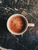 A steaming cup filled with frothy coffee sits on a dark, textured marble surface. The coffee shows a rich brown color with bubbles forming on the surface, suggesting a freshly poured and possibly creamy beverage.