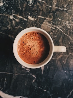 A steaming cup filled with frothy coffee sits on a dark, textured marble surface. The coffee shows a rich brown color with bubbles forming on the surface, suggesting a freshly poured and possibly creamy beverage.