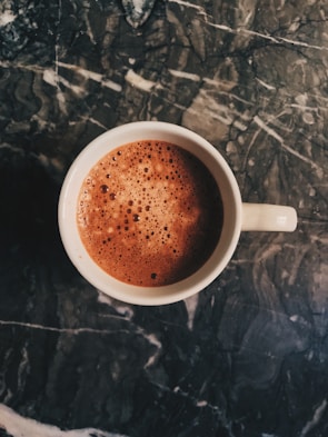 A steaming cup filled with frothy coffee sits on a dark, textured marble surface. The coffee shows a rich brown color with bubbles forming on the surface, suggesting a freshly poured and possibly creamy beverage.