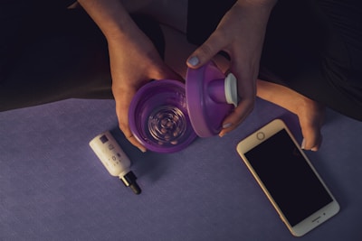 A person is sitting cross-legged on a yoga mat, holding a purple container with a white lid in their hands. A dropper bottle and a smartphone are placed next to them on the mat.