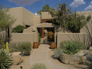 A Southwestern-style building with adobe-like walls surrounded by desert landscaping. There are two large clay pots flanking the entrance of a walkway that leads to a wooden door. Cacti, succulents, and desert shrubs are planted in a gravel garden, and a large rock is visible in the foreground. The sky is partially cloudy, and there are shadows cast by the plants and structure.