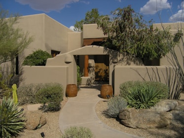 A Southwestern-style building with adobe-like walls surrounded by desert landscaping. There are two large clay pots flanking the entrance of a walkway that leads to a wooden door. Cacti, succulents, and desert shrubs are planted in a gravel garden, and a large rock is visible in the foreground. The sky is partially cloudy, and there are shadows cast by the plants and structure.