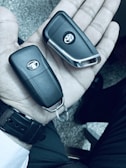 A hand holding two black car key fobs, each with a logo. The background is a textured surface, and the person is wearing a watch with a black strap.