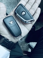 A hand holding two black car key fobs, each with a logo. The background is a textured surface, and the person is wearing a watch with a black strap.