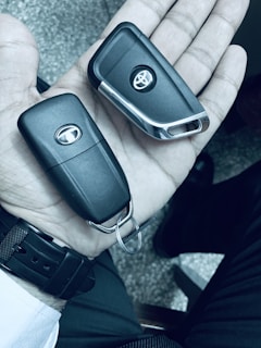 A hand holding two black car key fobs, each with a logo. The background is a textured surface, and the person is wearing a watch with a black strap.