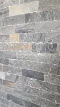 A wall composed of rectangular stone tiles with varying shades of gray and some beige accents. The surface has a rough, textured appearance with visible patterns and marks characteristic of natural stone.