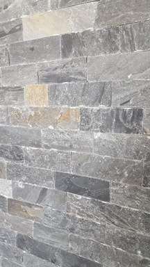 A wall composed of rectangular stone tiles with varying shades of gray and some beige accents. The surface has a rough, textured appearance with visible patterns and marks characteristic of natural stone.