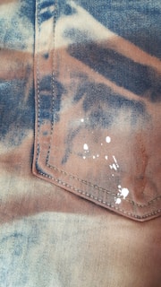 A close-up view of a piece of fabric with a pocket. The material appears to have a tie-dye pattern with brown and blue shades. There are small white paint splatters on the fabric, which add a textured element to the design.