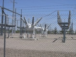 High-voltage electrical equipment is situated behind a chain-link fence topped with barbed wire. The area is a substation with large insulators and transformers, surrounded by gravel. Power lines and utility poles are visible in the background against a clear blue sky.