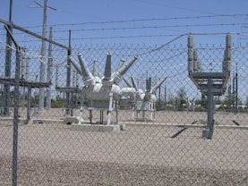 High-voltage electrical equipment is situated behind a chain-link fence topped with barbed wire. The area is a substation with large insulators and transformers, surrounded by gravel. Power lines and utility poles are visible in the background against a clear blue sky.