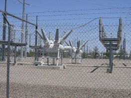 High-voltage electrical equipment is situated behind a chain-link fence topped with barbed wire. The area is a substation with large insulators and transformers, surrounded by gravel. Power lines and utility poles are visible in the background against a clear blue sky.