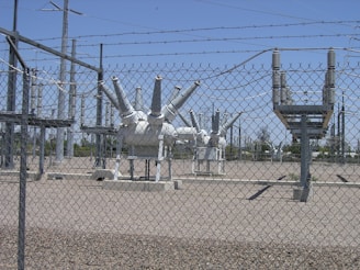 High-voltage electrical equipment is situated behind a chain-link fence topped with barbed wire. The area is a substation with large insulators and transformers, surrounded by gravel. Power lines and utility poles are visible in the background against a clear blue sky.