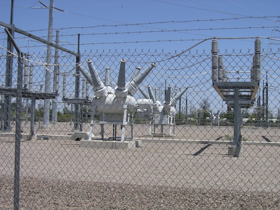 High-voltage electrical equipment is situated behind a chain-link fence topped with barbed wire. The area is a substation with large insulators and transformers, surrounded by gravel. Power lines and utility poles are visible in the background against a clear blue sky.