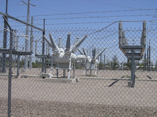 High-voltage electrical equipment is situated behind a chain-link fence topped with barbed wire. The area is a substation with large insulators and transformers, surrounded by gravel. Power lines and utility poles are visible in the background against a clear blue sky.
