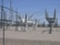 High-voltage electrical equipment is situated behind a chain-link fence topped with barbed wire. The area is a substation with large insulators and transformers, surrounded by gravel. Power lines and utility poles are visible in the background against a clear blue sky.