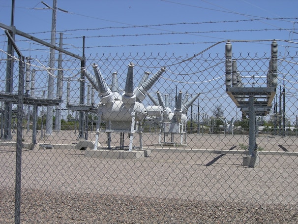 High-voltage electrical equipment is situated behind a chain-link fence topped with barbed wire. The area is a substation with large insulators and transformers, surrounded by gravel. Power lines and utility poles are visible in the background against a clear blue sky.