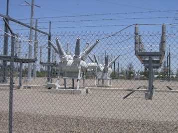High-voltage electrical equipment is situated behind a chain-link fence topped with barbed wire. The area is a substation with large insulators and transformers, surrounded by gravel. Power lines and utility poles are visible in the background against a clear blue sky.