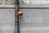 A copper drainpipe is mounted on a rough stone wall with a flared connector. The wall appears to be made of gray bricks that vary slightly in texture and color.