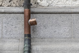 A copper drainpipe is mounted on a rough stone wall with a flared connector. The wall appears to be made of gray bricks that vary slightly in texture and color.
