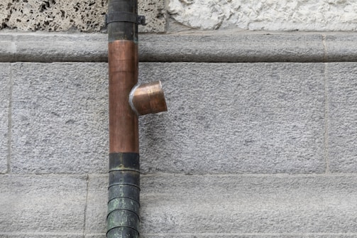 A copper drainpipe is mounted on a rough stone wall with a flared connector. The wall appears to be made of gray bricks that vary slightly in texture and color.
