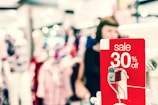 A bright and busy shopping environment with a prominent red sale sign indicating a 30% discount. The background is blurred, showing clothing displays and a person browsing, contributing to a lively atmosphere.