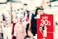 A bright and busy shopping environment with a prominent red sale sign indicating a 30% discount. The background is blurred, showing clothing displays and a person browsing, contributing to a lively atmosphere.