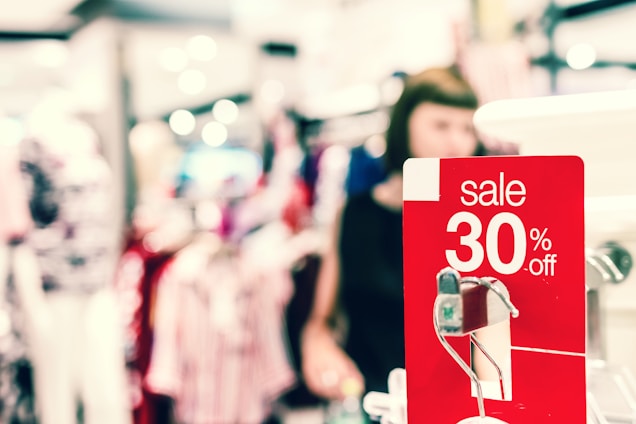 A bright and busy shopping environment with a prominent red sale sign indicating a 30% discount. The background is blurred, showing clothing displays and a person browsing, contributing to a lively atmosphere.