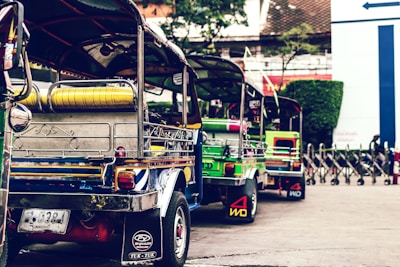 A row of vibrant, colorful tuk-tuks is parked along a street. These three-wheeled vehicles are adorned with bright colors and various decorative elements, such as metal detailing and logos on the back panels. They are aligned closely together, suggesting a busy, possibly urban setting. In the background, there are blurred elements of greenery and a metal barrier, indicating an outdoor environment.