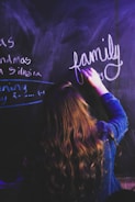 A person with long, wavy hair is writing the word 'family' in white chalk on a dark chalkboard. The surrounding area shows partially visible writing in chalk in different styles and directions. The person's back is turned to the viewer and they are wearing a denim jacket.