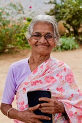 An elderly woman with gray hair and glasses stands outdoors, smiling warmly. She wears a floral-patterned saree and holds a book close to her chest. The background features lush green foliage and a sandy area.