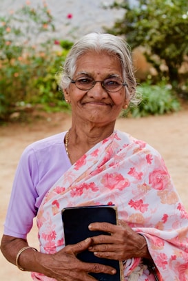 An elderly woman with gray hair and glasses stands outdoors, smiling warmly. She wears a floral-patterned saree and holds a book close to her chest. The background features lush green foliage and a sandy area.