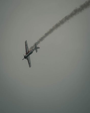 An aircraft is captured mid-flight, performing an acrobatic maneuver with a trail of smoke following behind against a muted sky. The vehicle is angled as if turning sharply, suggesting dynamic movement.