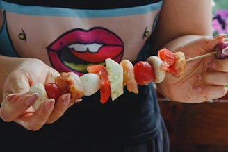 A person holding a skewer loaded with various ingredients including red peppers, tomatoes, onions, and chunks of meat. The person is wearing a shirt with a stylized graphic of lips. The image has a casual, homemade feel with a focus on preparing food.