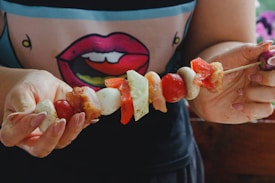 A person holding a skewer loaded with various ingredients including red peppers, tomatoes, onions, and chunks of meat. The person is wearing a shirt with a stylized graphic of lips. The image has a casual, homemade feel with a focus on preparing food.