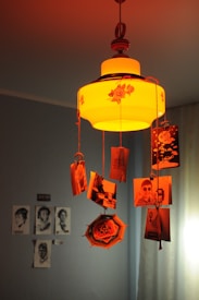 An ornate, vintage-style ceiling lamp with floral designs casts a warm, orange glow. Photographs are attached to strings hanging from the lamp, creating a nostalgic and personalized decor. Shadows and light interplay on the surrounding walls.