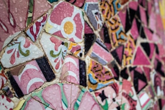 A vibrant mosaic art piece made up of various colored ceramic tiles, featuring intricate patterns and floral designs. The tiles are predominantly pink with accents of yellow, green, and black, and they appear glossy with water droplets on the surface.