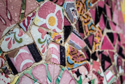A vibrant mosaic art piece made up of various colored ceramic tiles, featuring intricate patterns and floral designs. The tiles are predominantly pink with accents of yellow, green, and black, and they appear glossy with water droplets on the surface.