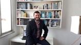 A man is sitting on the edge of a white desk in front of a fully stocked bookshelf. He is wearing a dark blazer over a maroon shirt, and is smiling at the camera. A closed laptop and a desk lamp are visible on the desk. A framed photograph and various books in different colors are neatly arranged on the shelves.