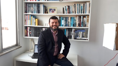 A man is sitting on the edge of a white desk in front of a fully stocked bookshelf. He is wearing a dark blazer over a maroon shirt, and is smiling at the camera. A closed laptop and a desk lamp are visible on the desk. A framed photograph and various books in different colors are neatly arranged on the shelves.