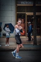 A person wearing a sporty outfit, including a headband, tight-fitting tank top, and shorts, is walking on a street. They appear to be gesturing playfully with their hands. In the background, a store with advertisements, and people, some of whom are holding umbrellas, are visible standing on the sidewalk.