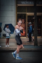 A person wearing a sporty outfit, including a headband, tight-fitting tank top, and shorts, is walking on a street. They appear to be gesturing playfully with their hands. In the background, a store with advertisements, and people, some of whom are holding umbrellas, are visible standing on the sidewalk.