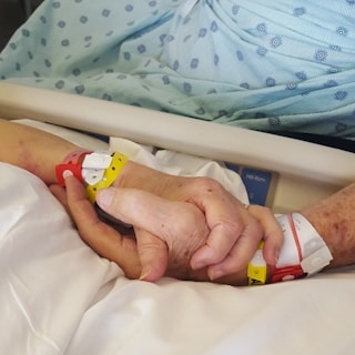 Two hands are clasped together, both wearing multiple hospital wristbands. The background includes a hospital bed with a patterned hospital gown visible.