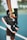 A person holding a pair of sneakers with black, white, and orange accents, standing on a blue tennis court. The focus is on the shoes, with the person slightly out of frame, wearing black shorts with a neon stripe and a white shirt.