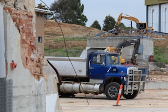 A construction site with a blue truck parked prominently in the foreground. The scene includes earthmoving equipment like an excavator in the background. There is a partially demolished brick wall on the left side, adding to the industrial atmosphere. The setting appears under a cloudy sky with trees in the distance.